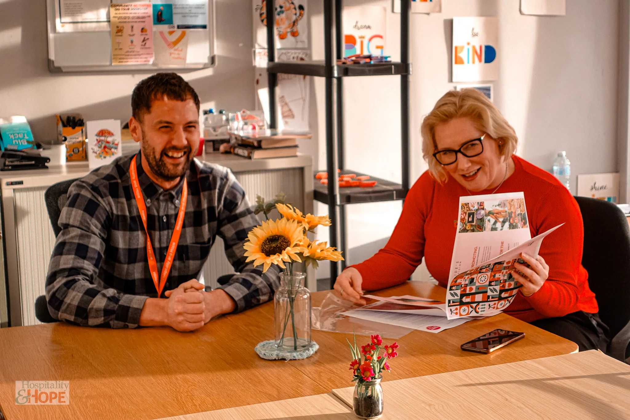 Office scene with two people at a meeting table; man in plaid shirt and orange lanyard smiles while a woman in red reads a colorful magazine, sunflowers on the table.
