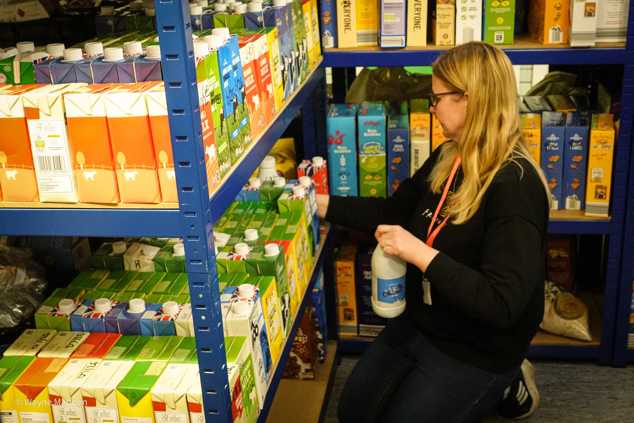 Woman kneeling and stocking juice cartons on blue metal shelves in a store aisle.