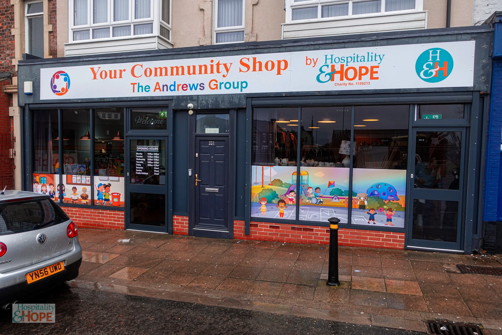 Storefront sign reading 'Your Community Shop' with 'The Andrews Group' and 'Hospitality & Hope' logo on a gray facade, large shop windows with colorful child-friendly graphics